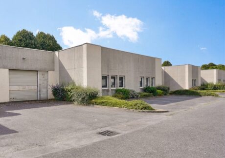Immeuble commercial beige de plain-pied, doté de grandes fenêtres, d'une porte de garage, d'arbustes bien entretenus et d'un parking vide, sous un ciel bleu parsemé de nuages.