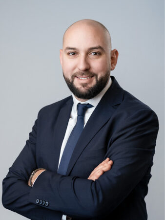 Un homme en costume-cravate se tient debout, les bras croisés, souriant à l'appareil photo sur un fond gris clair uni.