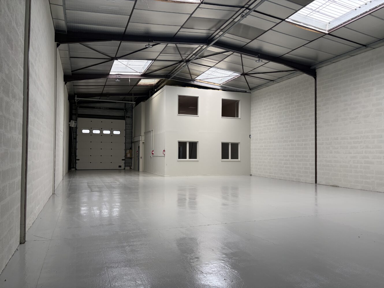 Intérieur d'un entrepôt vide aux murs blancs, avec un sol en béton poli, un haut plafond et un petit bureau situé près d'une porte de garage fermée.