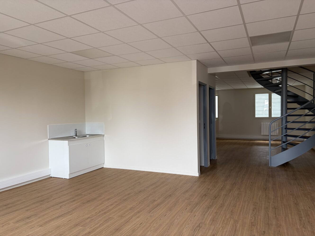 Chambre non meublée, avec un petit meuble-lavabo blanc, des murs clairs, du parquet et un escalier en colimaçon métallique en arrière-plan.
