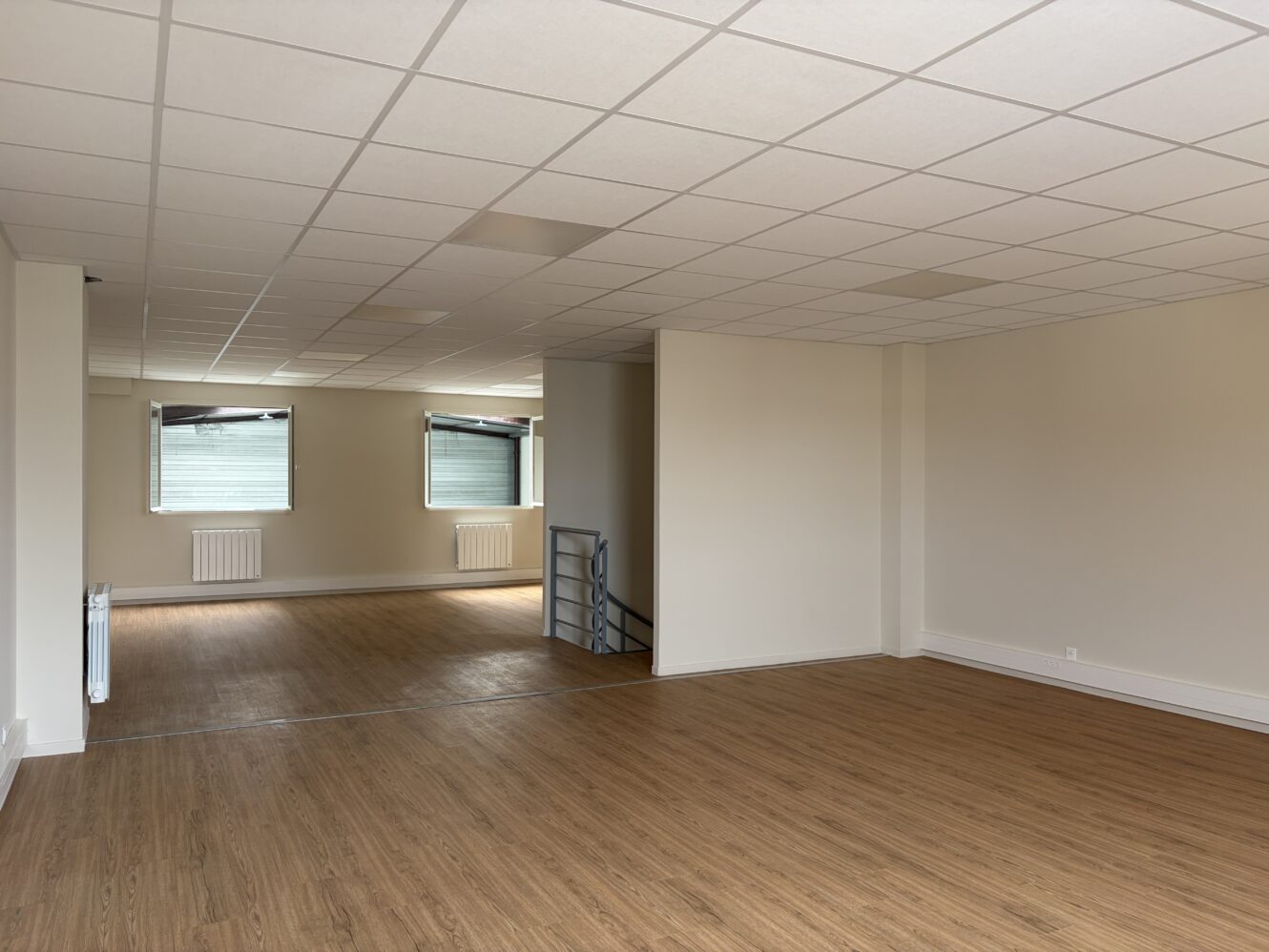Pièce vide aux murs blancs, dotée d'un faux plafond, d'un parquet, de deux fenêtres et d'une petite balustrade métallique près d'une marche menant à un autre espace.