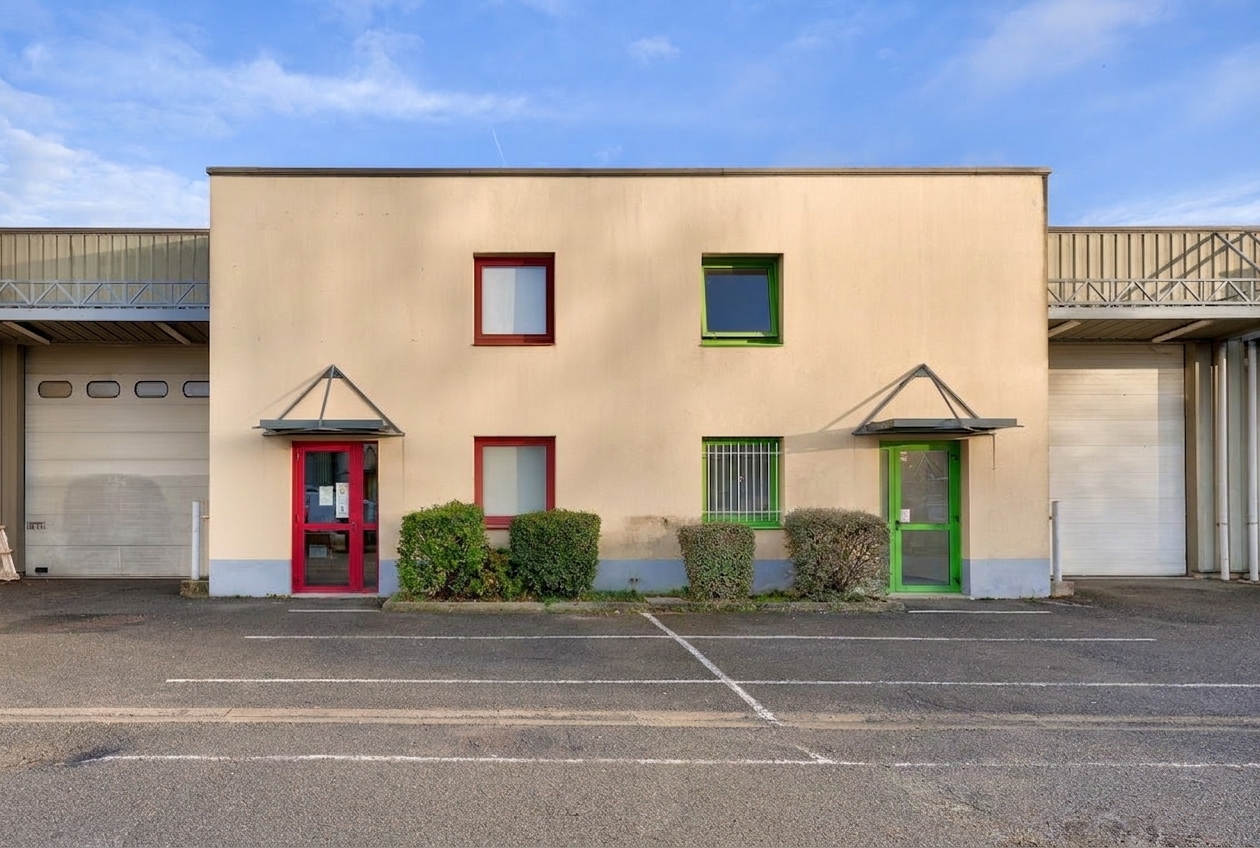 Un bâtiment industriel beige doté de deux portes et fenêtres, dont l'une est encadrée de rouge et l'autre de vert, avec des buissons devant et des places de parking vides.
