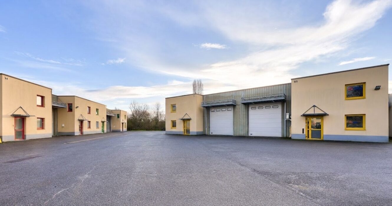 Un parking pavé situé devant plusieurs bâtiments industriels jaunes, dotés de grandes portes de garage et de fenêtres carrées, sous un ciel partiellement nuageux.