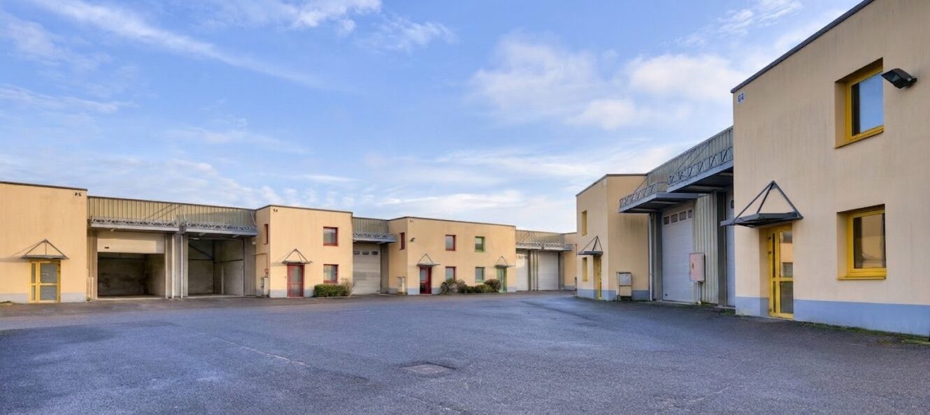 Un parking pavé devant plusieurs entrepôts industriels de couleur beige, aux fenêtres et portes encadrées de jaune, sous un ciel partiellement nuageux.