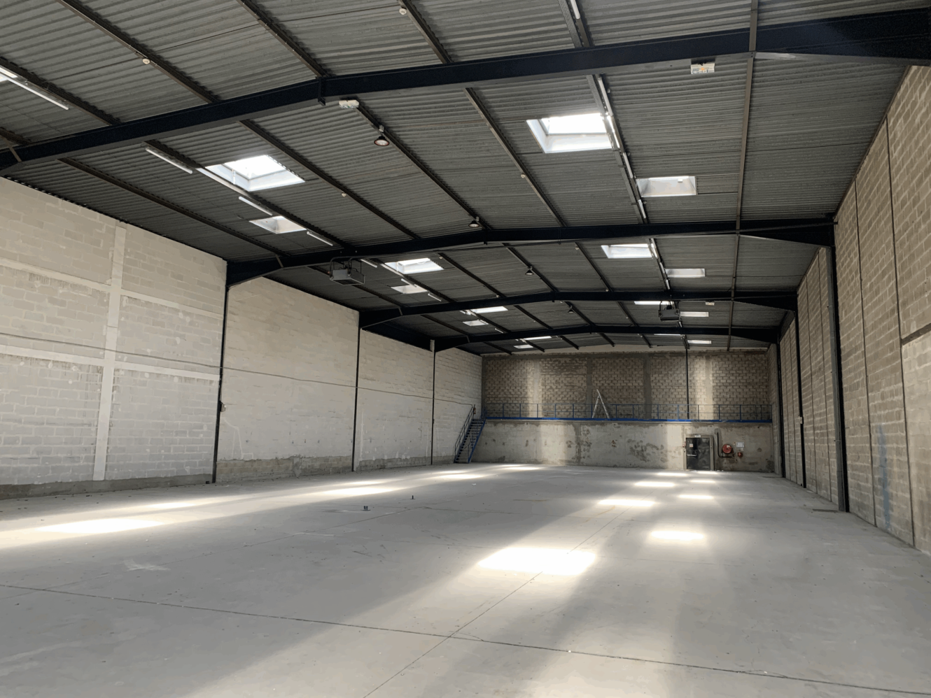 Intérieur d'un entrepôt vide avec de hauts plafonds, des poutres métalliques apparentes, des puits de lumière, des murs en béton et une mezzanine à l'extrémité.