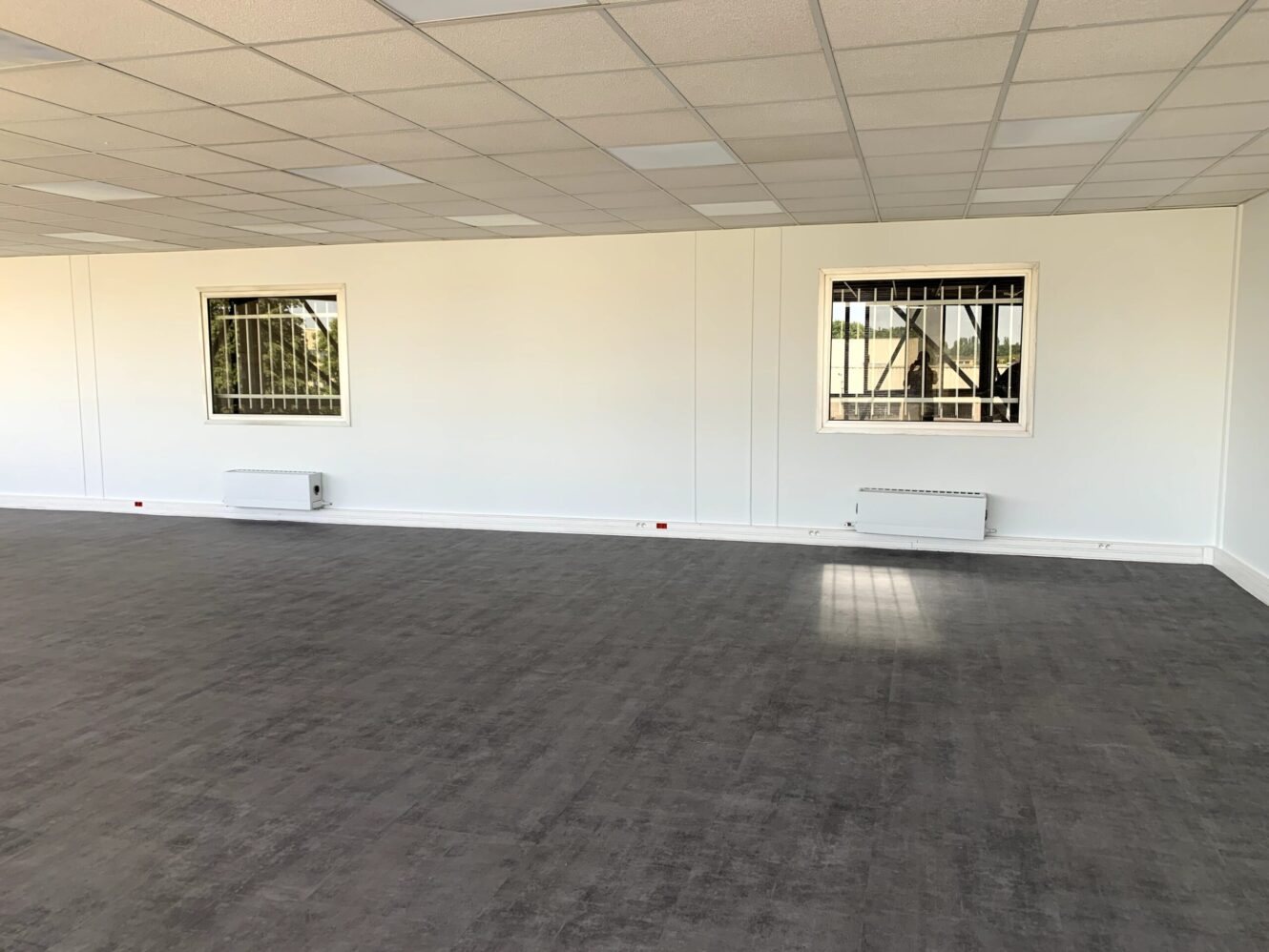 Une pièce vide avec un sol gris, des murs blancs, deux fenêtres grillagées et deux radiateurs muraux sous les fenêtres.