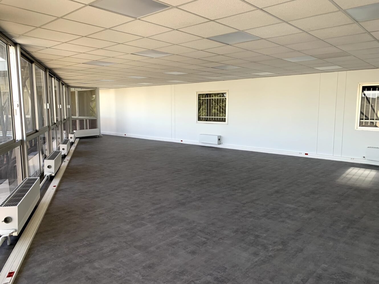 Un grand espace de bureau vide avec un sol gris, des murs blancs, un plafond quadrillé et des fenêtres le long d'un côté qui laissent entrer la lumière naturelle.