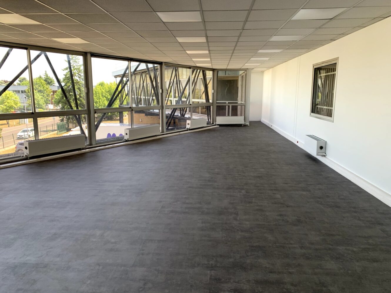 Un espace de bureau vide avec de grandes fenêtres, des murs blancs et un sol sombre, avec des unités de chauffage le long des fenêtres et une vue sur les arbres et les bâtiments à l'extérieur.