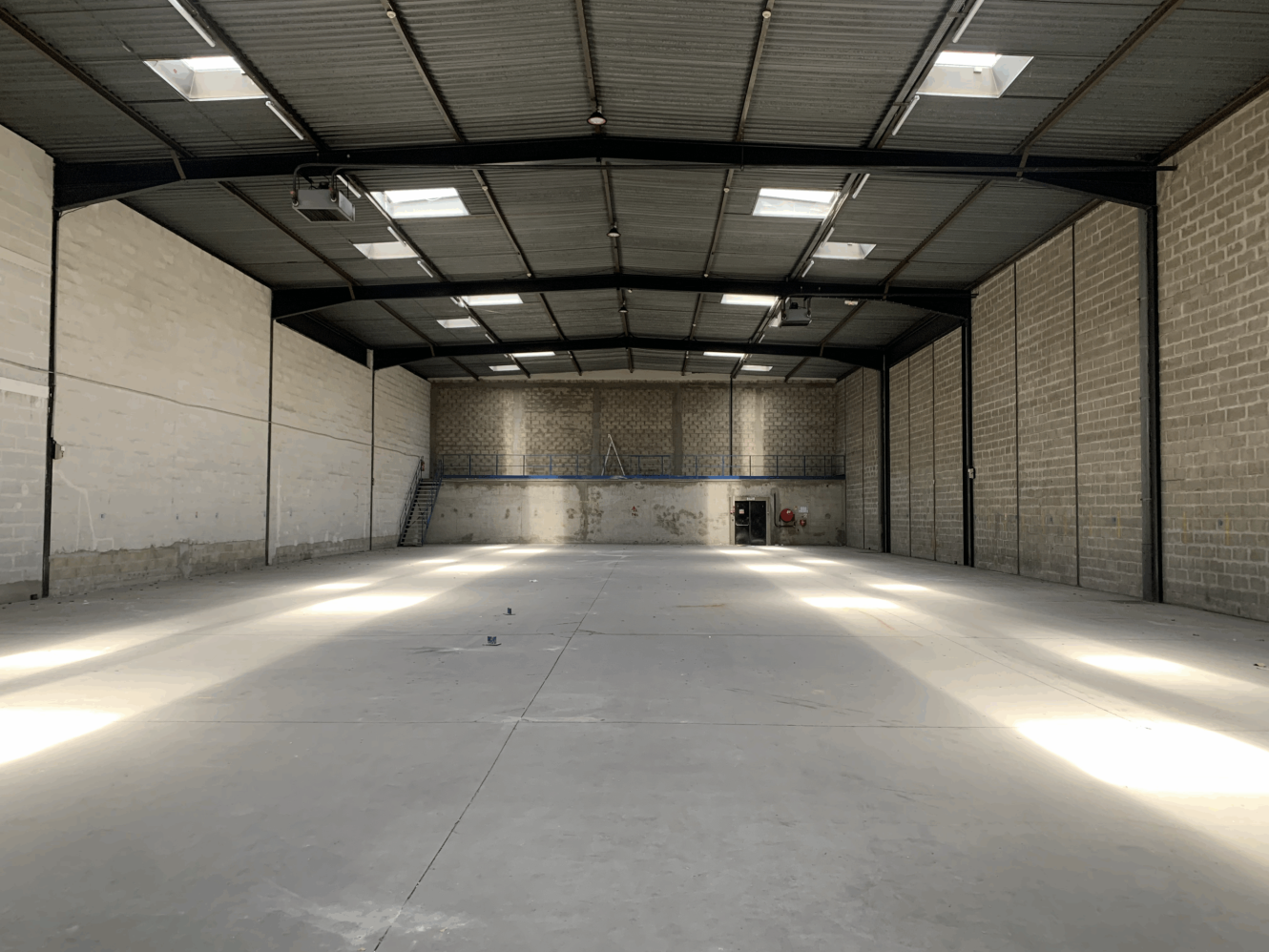 Entrepôt industriel vide avec de hauts plafonds, des sols en béton, des murs en briques et des lucarnes laissant entrer la lumière naturelle. Un escalier est visible à l'extrémité.