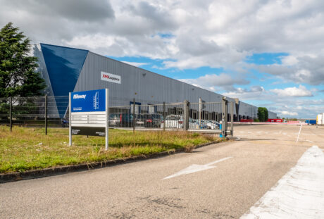 Un grand entrepôt de XPO Logistics avec une aire de stationnement clôturée, des véhicules garés, un panneau d'entrée et un ciel nuageux au-dessus de la tête.