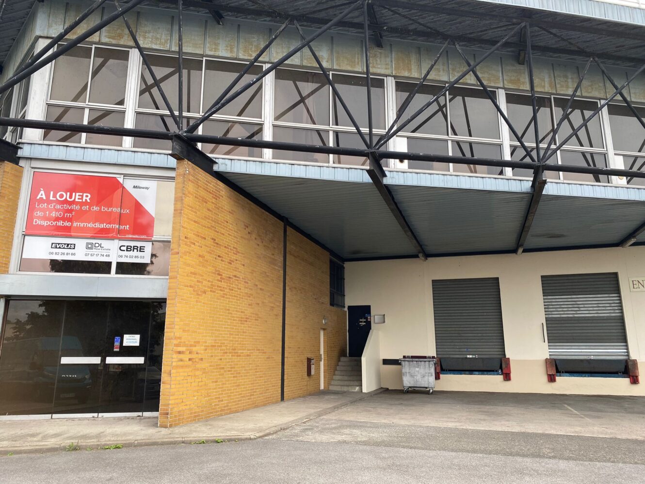Bâtiment industriel avec panneau de location de bureaux en français, montrant des quais de chargement, une benne à ordures et des portes d'entrée sous un auvent en métal.