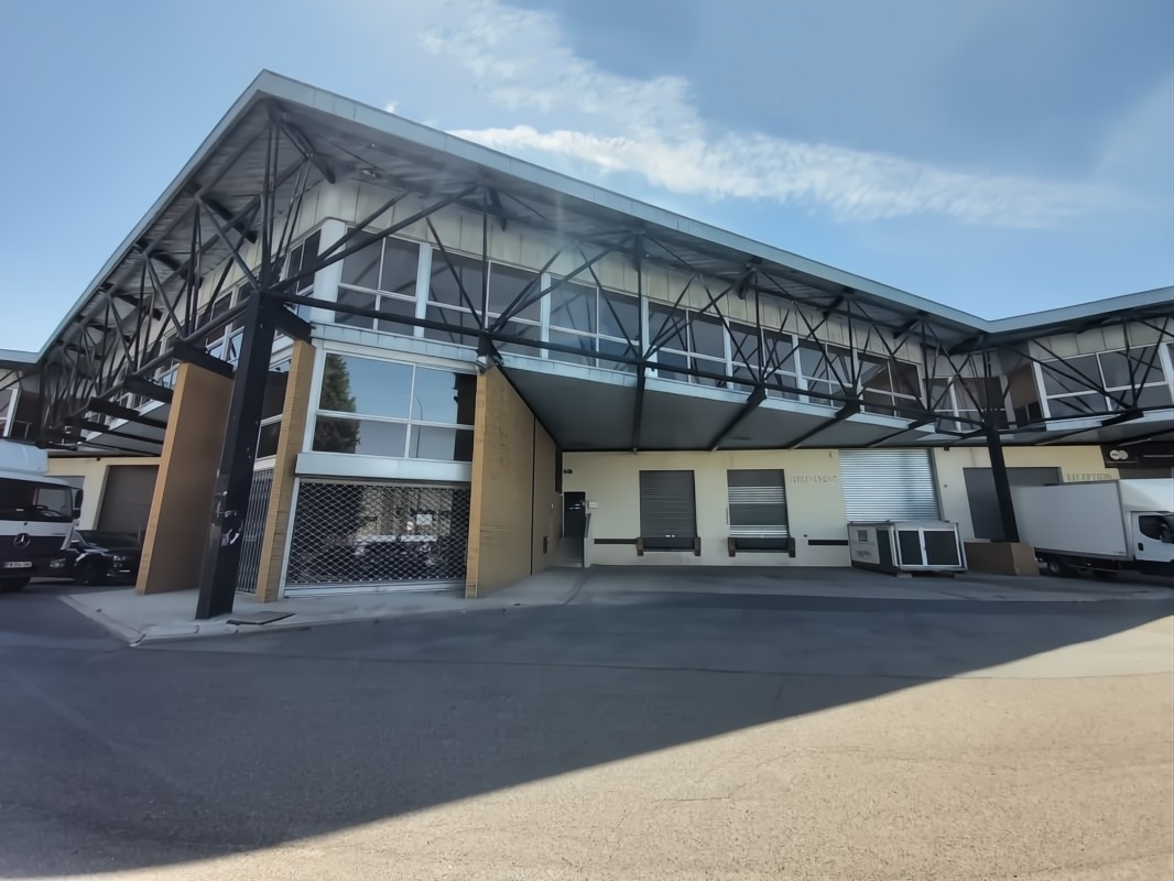 Un bâtiment industriel moderne avec de grandes fenêtres, des poutres métalliques, des quais de chargement et plusieurs camions de livraison garés à l'extérieur par une journée ensoleillée.