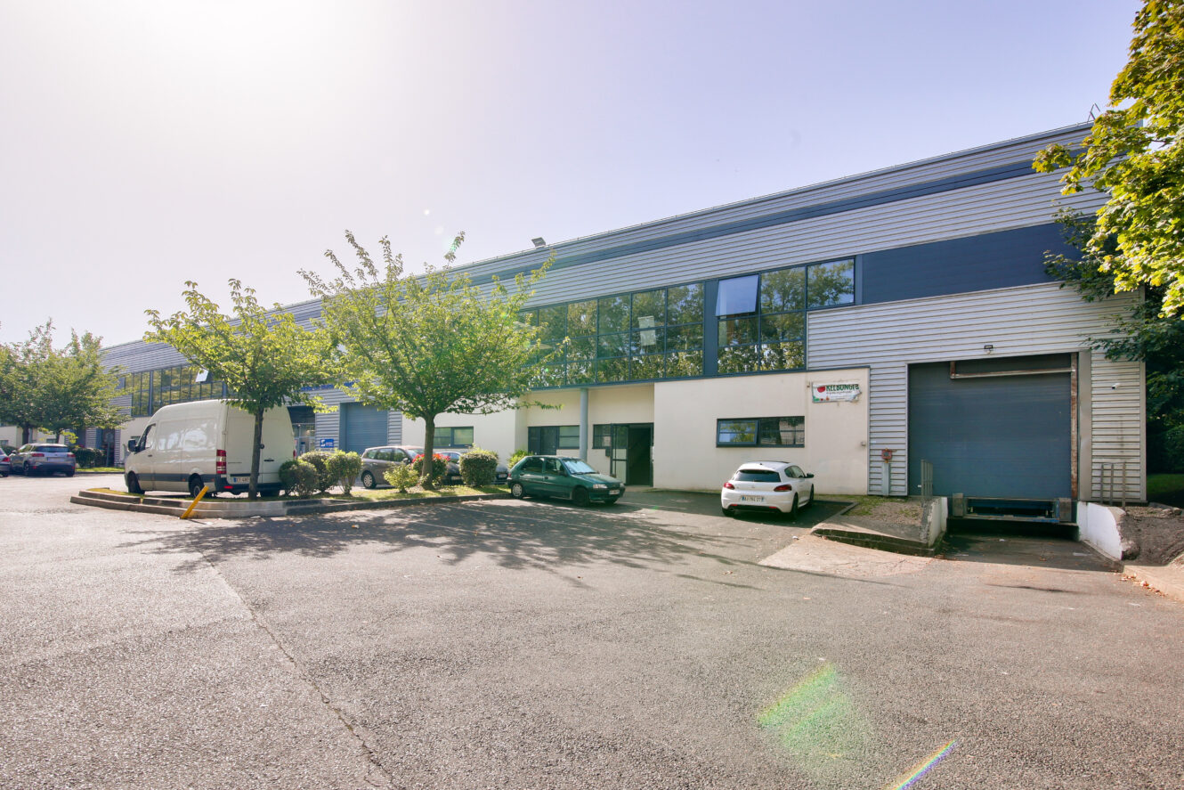 Un entrepôt commercial avec un quai de chargement, plusieurs véhicules garés, des arbres et un temps clair et ensoleillé.