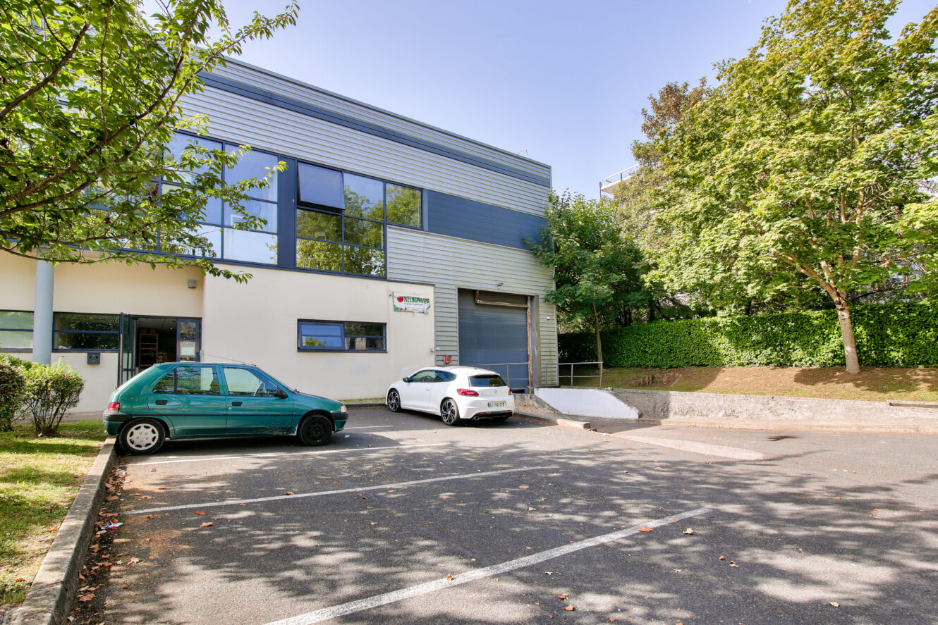 Un bâtiment commercial moderne avec de grandes fenêtres, une porte de garage, deux voitures garées et des arbres environnants par une journée ensoleillée.