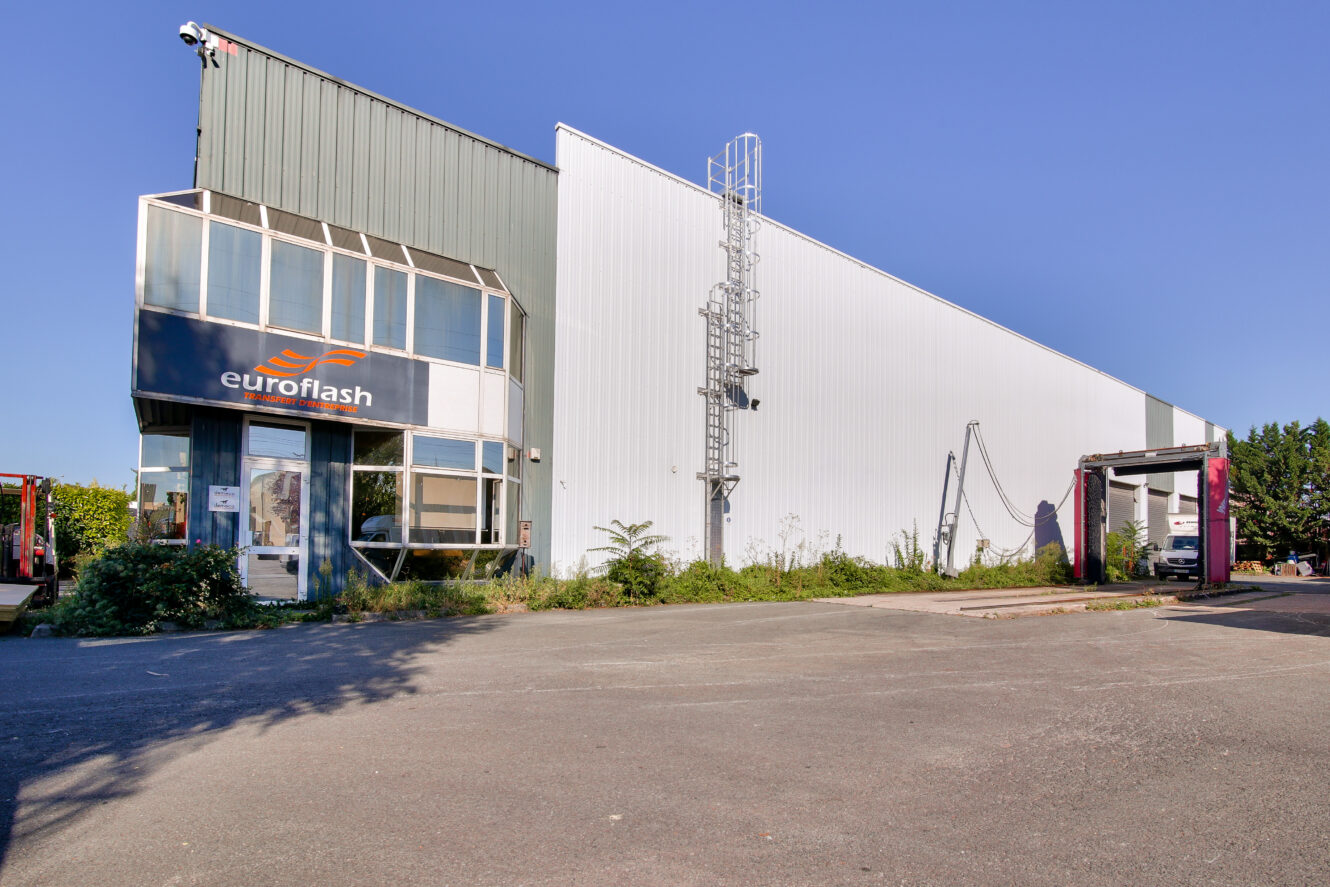 Un grand bâtiment industriel avec une enseigne euroflash, une entrée en verre, un extérieur en métal et une station de lavage adjacente sous un ciel bleu clair.