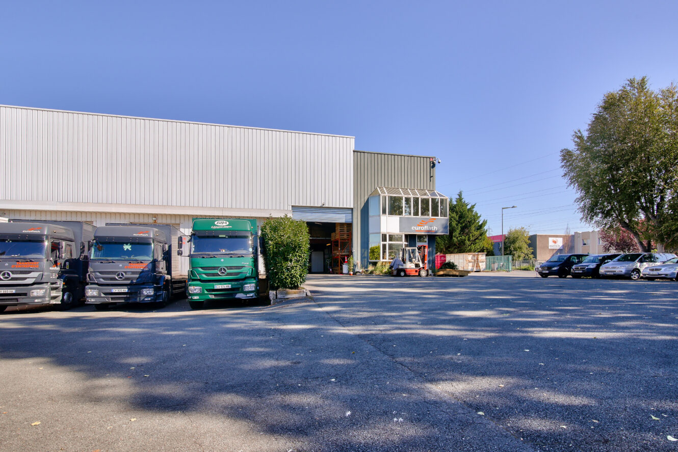 Plusieurs camions sont garés à l'extérieur d'un grand entrepôt industriel dont l'entrée est vitrée, sous un ciel dégagé.