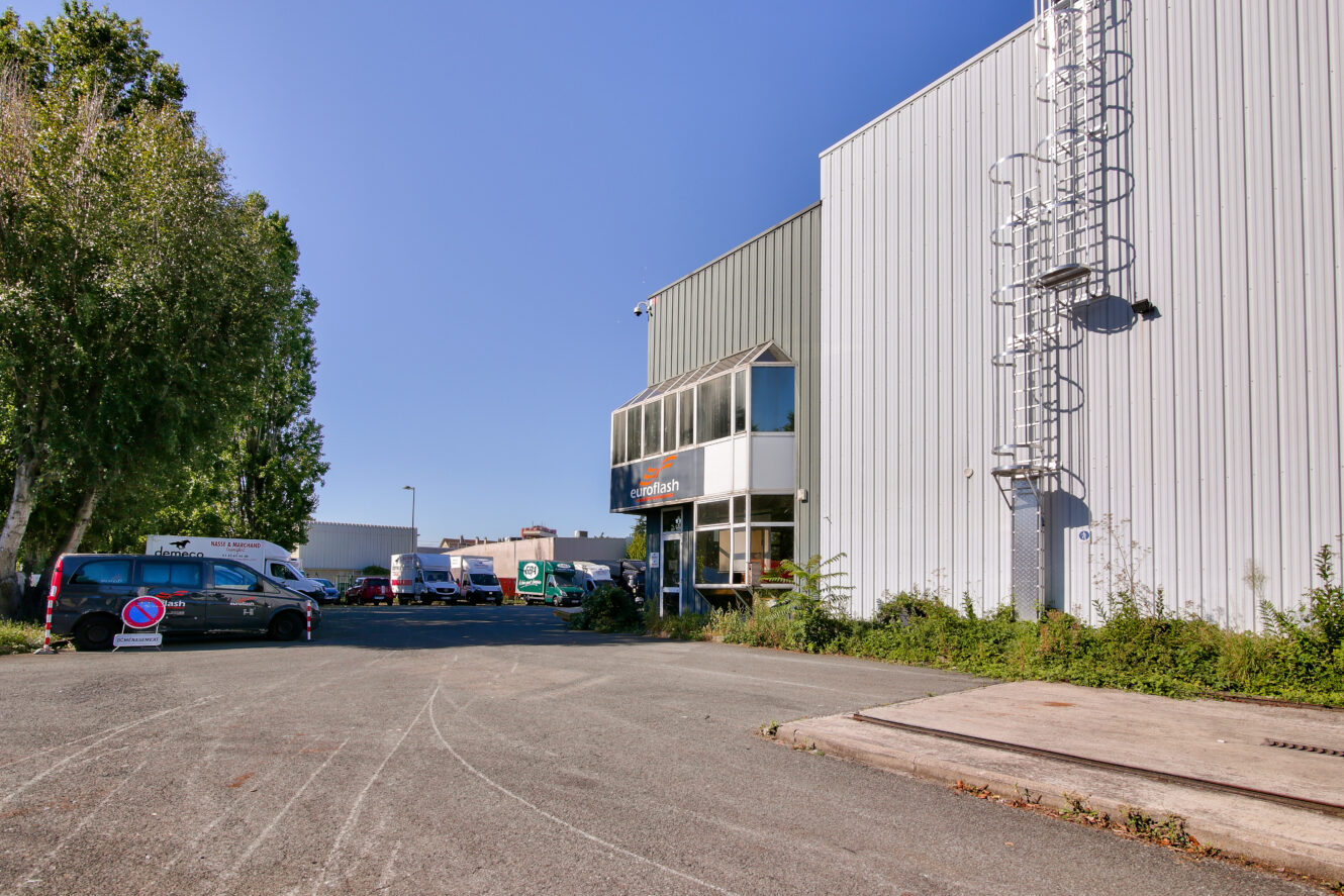 Un entrepôt commercial avec un extérieur métallique, des camionnettes garées et une petite entrée de bureau par une journée ensoleillée.