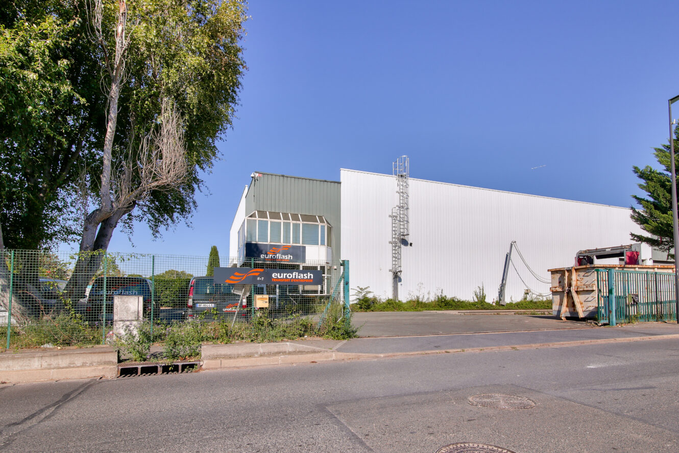 Un bâtiment industriel blanc avec un panneau euroflash, entouré d'une clôture métallique, de verdure et de matériaux de construction, sous un ciel bleu clair.