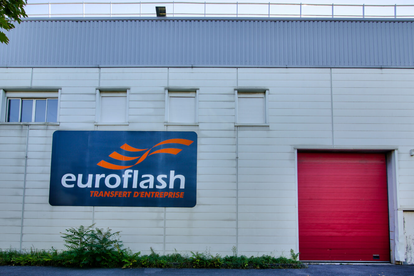 Vue extérieure d'un bâtiment avec une grande enseigne euroflash TRANSFERT DENTREPRISE et une porte de garage rouge fermée.