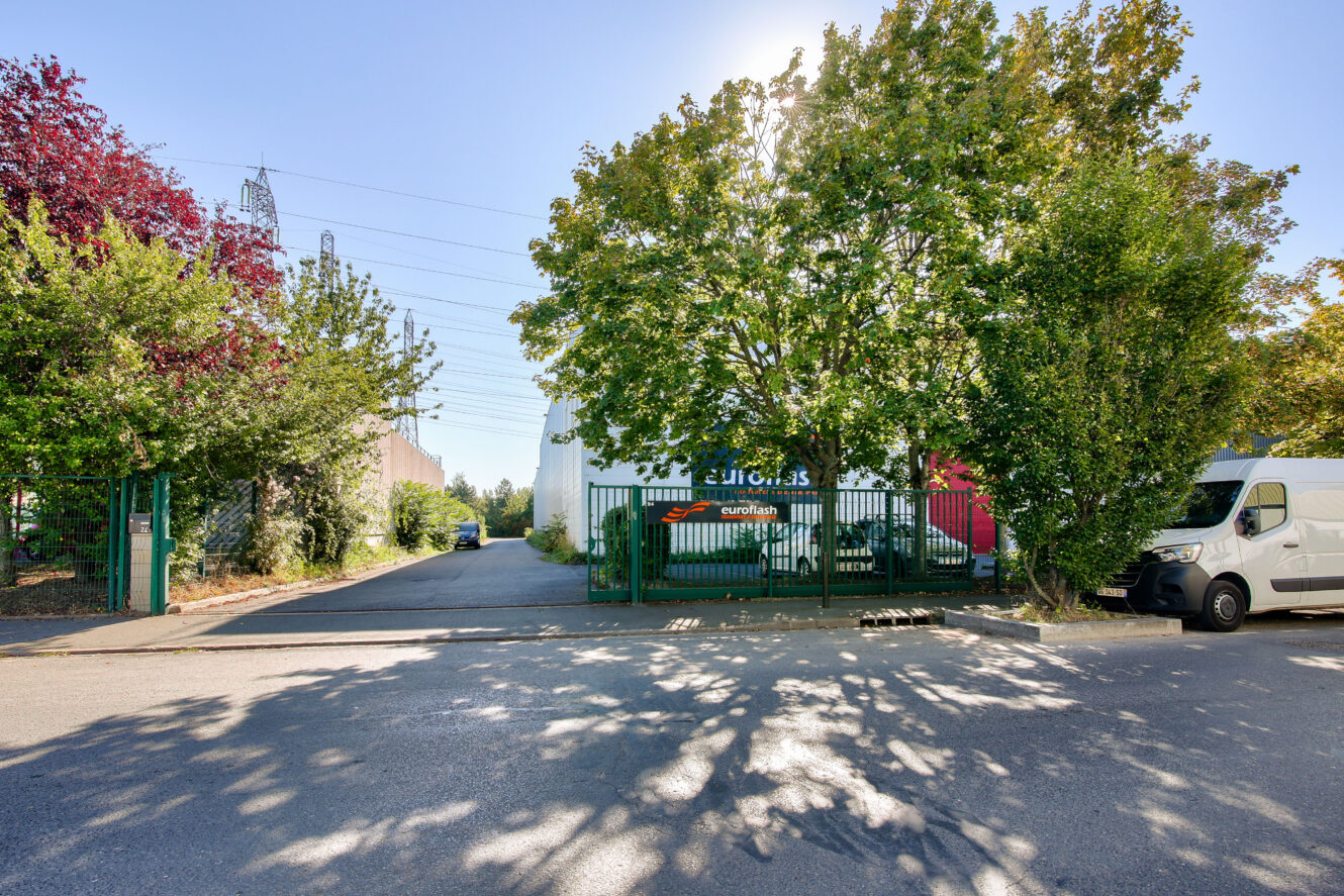 Un portail métallique vert partiellement masqué par des arbres mène à une allée pavée à côté d'un bâtiment blanc ; une camionnette blanche est garée sur le côté droit.