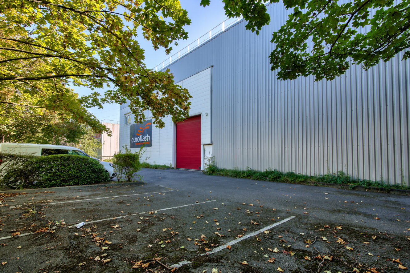 Un grand bâtiment industriel avec une porte roulante rouge et un panneau Eurotech, bordé d'arbres et d'un parking vide avec des feuilles éparses.