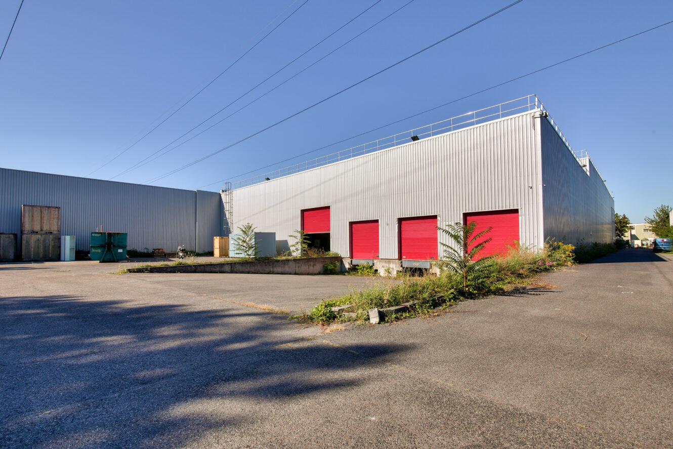 Entrepôt industriel avec un bardage métallique gris et quatre portes roulantes rouges, entouré d'un terrain pavé et d'une végétation éparse, sous un ciel bleu clair.