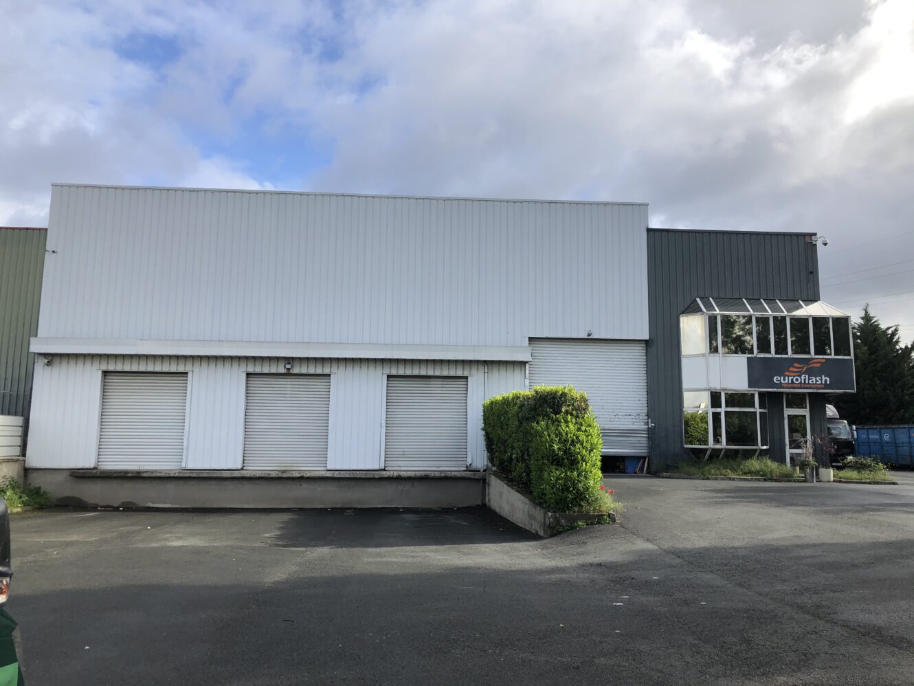 Bâtiment industriel avec trois volets métalliques fermés, une porte roulante et une entrée en verre étiquetée euroflash par temps nuageux.