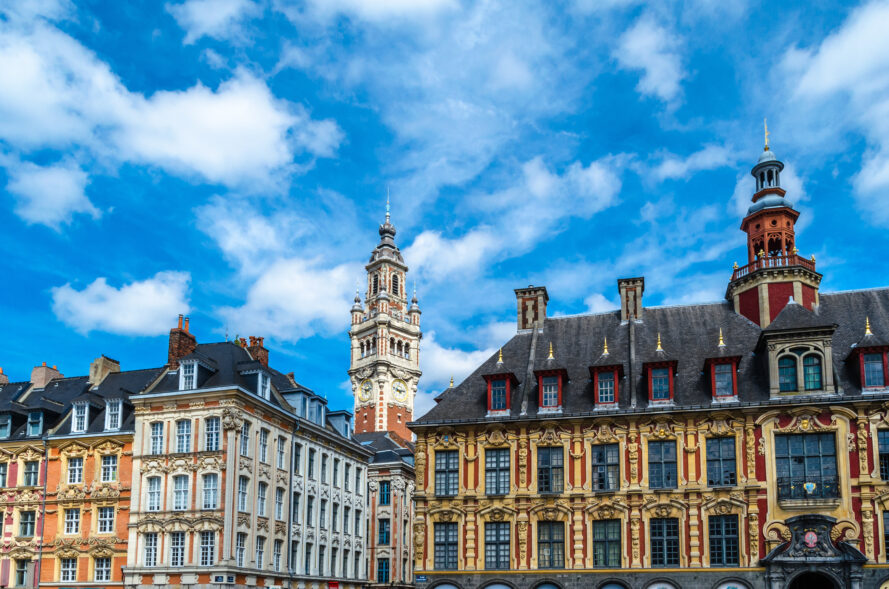 Bâtiments historiques aux façades ornées et tour d'horloge centrale sous un ciel bleu partiellement nuageux dans une ville européenne.