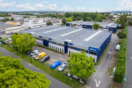 Un grand entrepôt industriel avec un toit gris et des accents bleus, entouré d'arbres, de véhicules en stationnement et de bâtiments adjacents, vu de dessus.