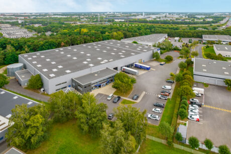 Vue aérienne d'un grand entrepôt industriel avec des camions aux quais de chargement et des voitures dans le parking, entouré d'arbres et de bâtiments industriels voisins.