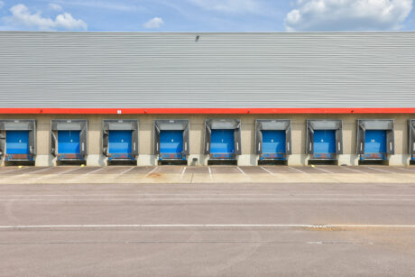 Une rangée de neuf quais de chargement vides avec des portes bleues fermées dans un entrepôt de distribution sous un ciel partiellement nuageux.