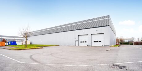 Un grand entrepôt industriel moderne avec deux portes de quai de chargement, un arbre, un parking et un ciel bleu en arrière-plan.