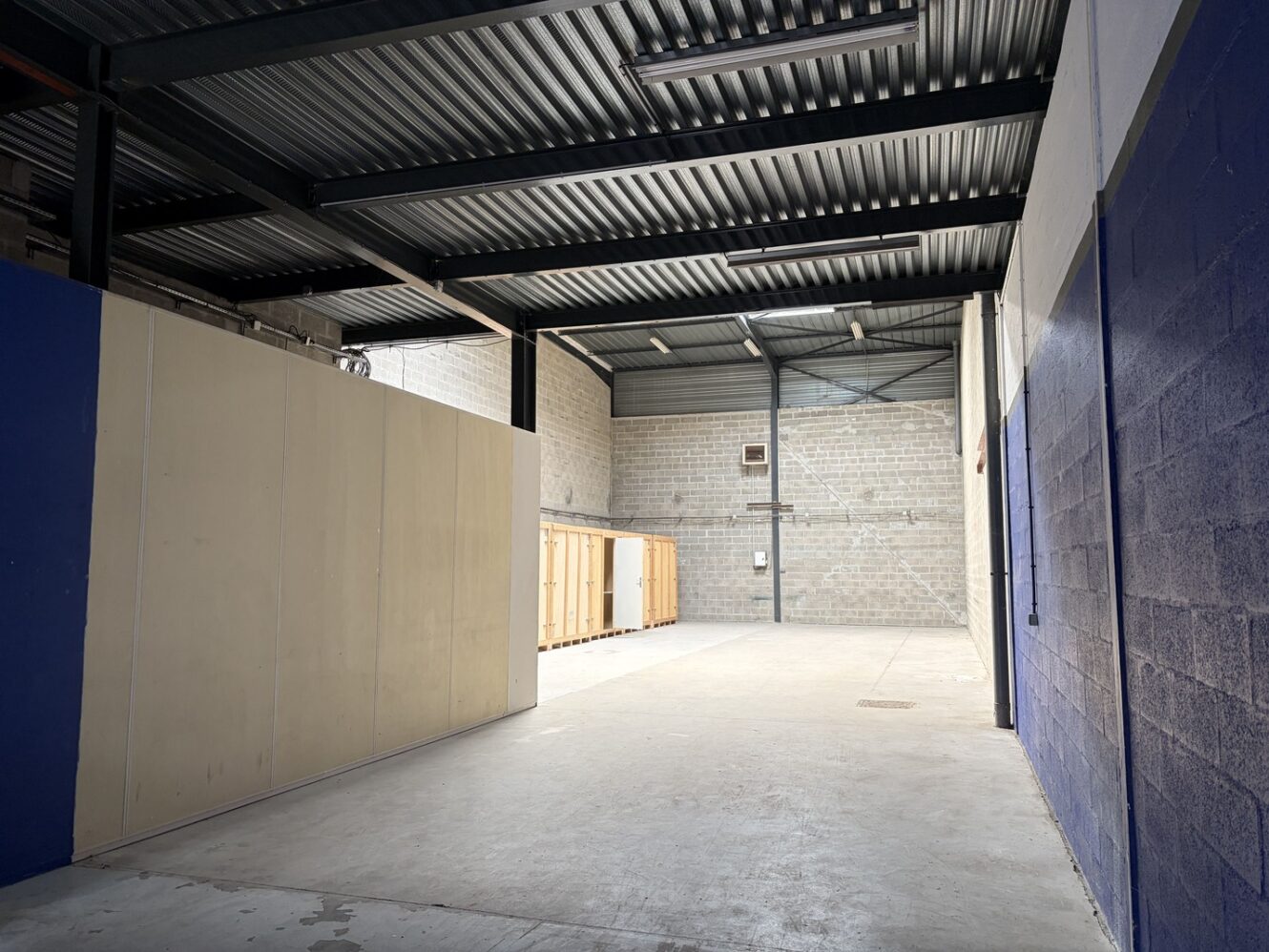 Intérieur d'un entrepôt vide avec un sol en béton, des murs bruts, des poutres métalliques au plafond et des rayonnages en bois le long du mur du fond.