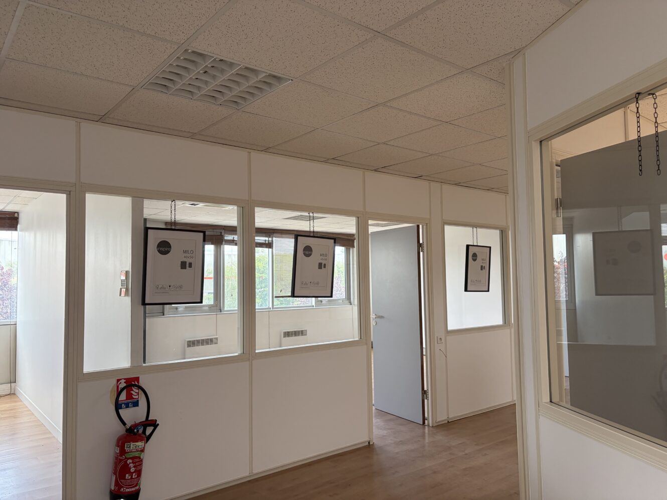 Un couloir dans un immeuble de bureaux, avec des murs blancs, des fenêtres vitrées, des certificats encadrés, un extincteur et une porte entrouverte.