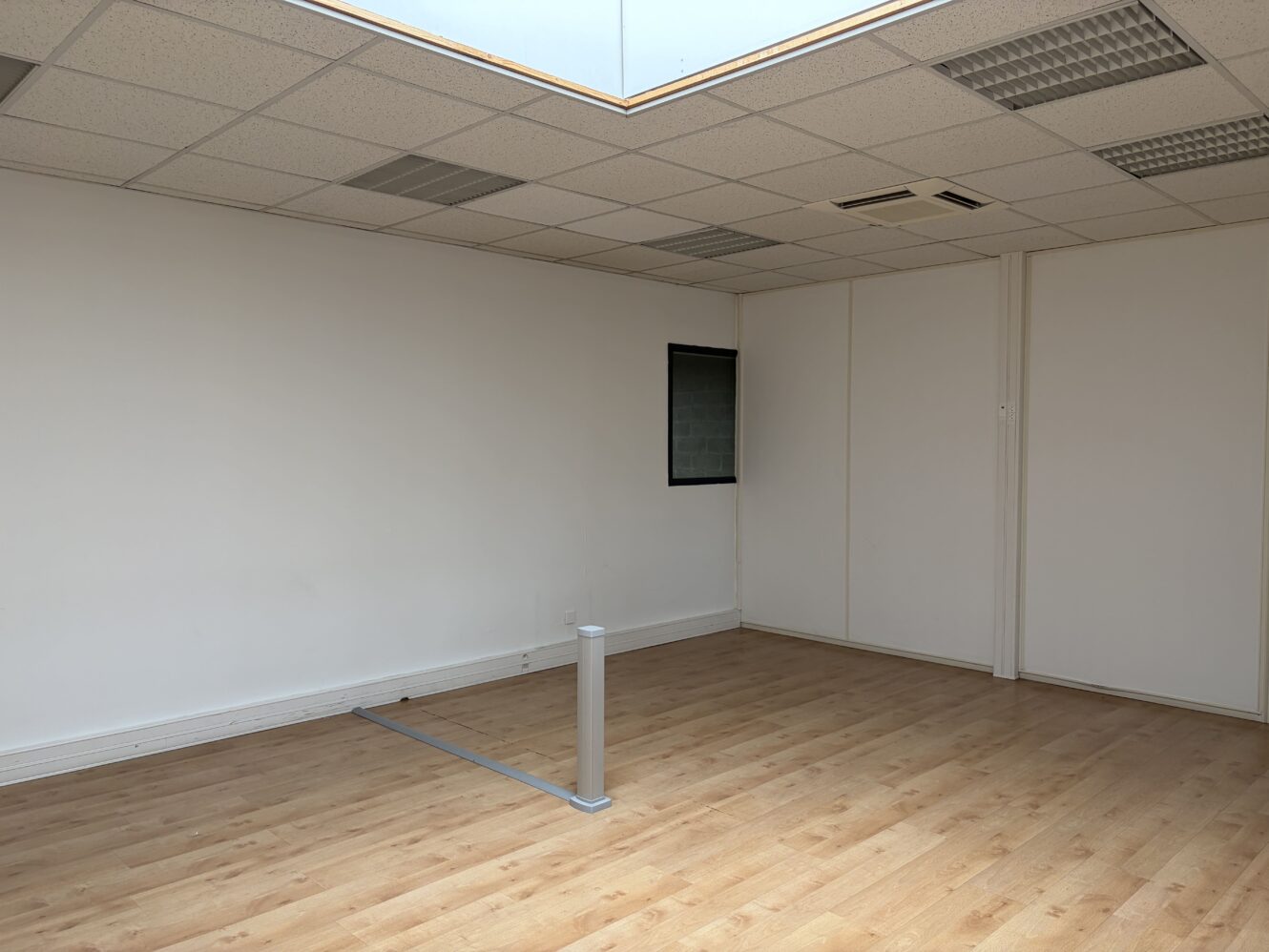 Bureau vide avec des murs blancs, du parquet, une petite fenêtre, des plafonniers et une prise de courant au sol.