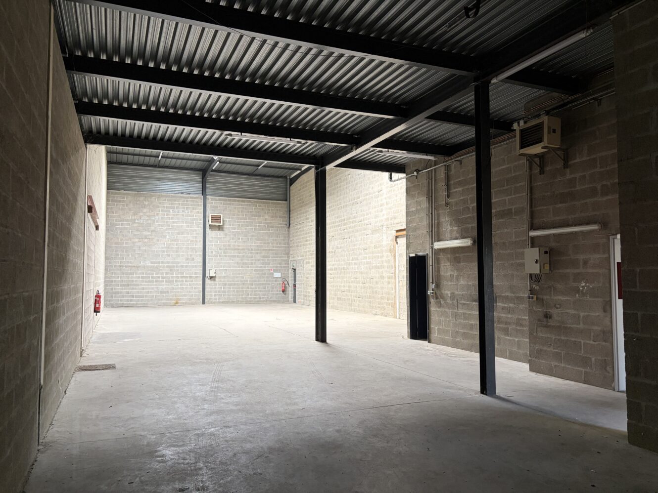Intérieur d'un entrepôt industriel vide, avec des murs en béton apparent, un plafond métallique, des poutres de soutien et des tableaux électriques ; lumière du soleil venant de la gauche.