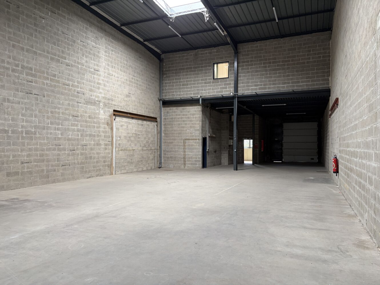 Entrepôt industriel vide avec sol en béton, grande hauteur sous plafond, lucarne, petite fenêtre, extincteur et grande porte de garage à l'arrière.