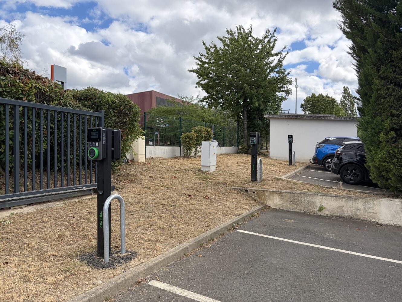 Des bornes de recharge pour véhicules électriques dans un parking, à côté de voitures garées, d'une clôture et d'arbres, sous un ciel nuageux.