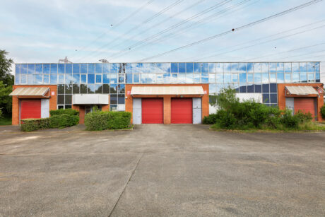 Un grand bâtiment industriel avec des fenêtres en verre miroir, trois portes de garage rouges et un terrain pavé vide devant. Des buissons et de la verdure sont visibles autour du bâtiment.