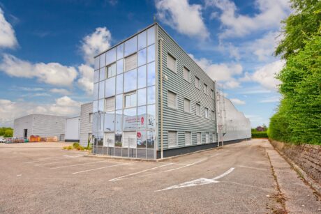 Un bâtiment industriel moderne de trois étages avec un revêtement en verre et en métal, adjacent à un parking vide, sous un ciel partiellement nuageux.