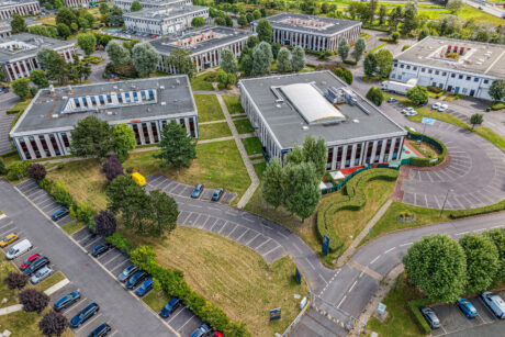 Vue aérienne d'un parc d'activités avec de nombreux immeubles de bureaux, des parkings avec des voitures, des pelouses vertes et des allées bordées d'arbres.