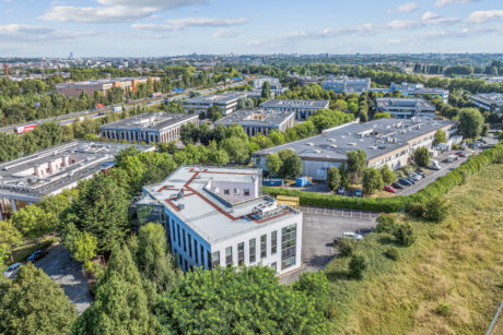 Vue aérienne d'un parc d'affaires comprenant plusieurs immeubles de bureaux modernes entourés d'arbres et de parkings, avec un paysage urbain en arrière-plan.