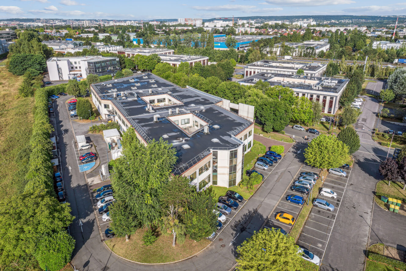 Vue aérienne d'immeubles de bureaux entourés d'arbres et de parkings avec plusieurs voitures garées, dans un quartier d'affaires urbain par temps clair.