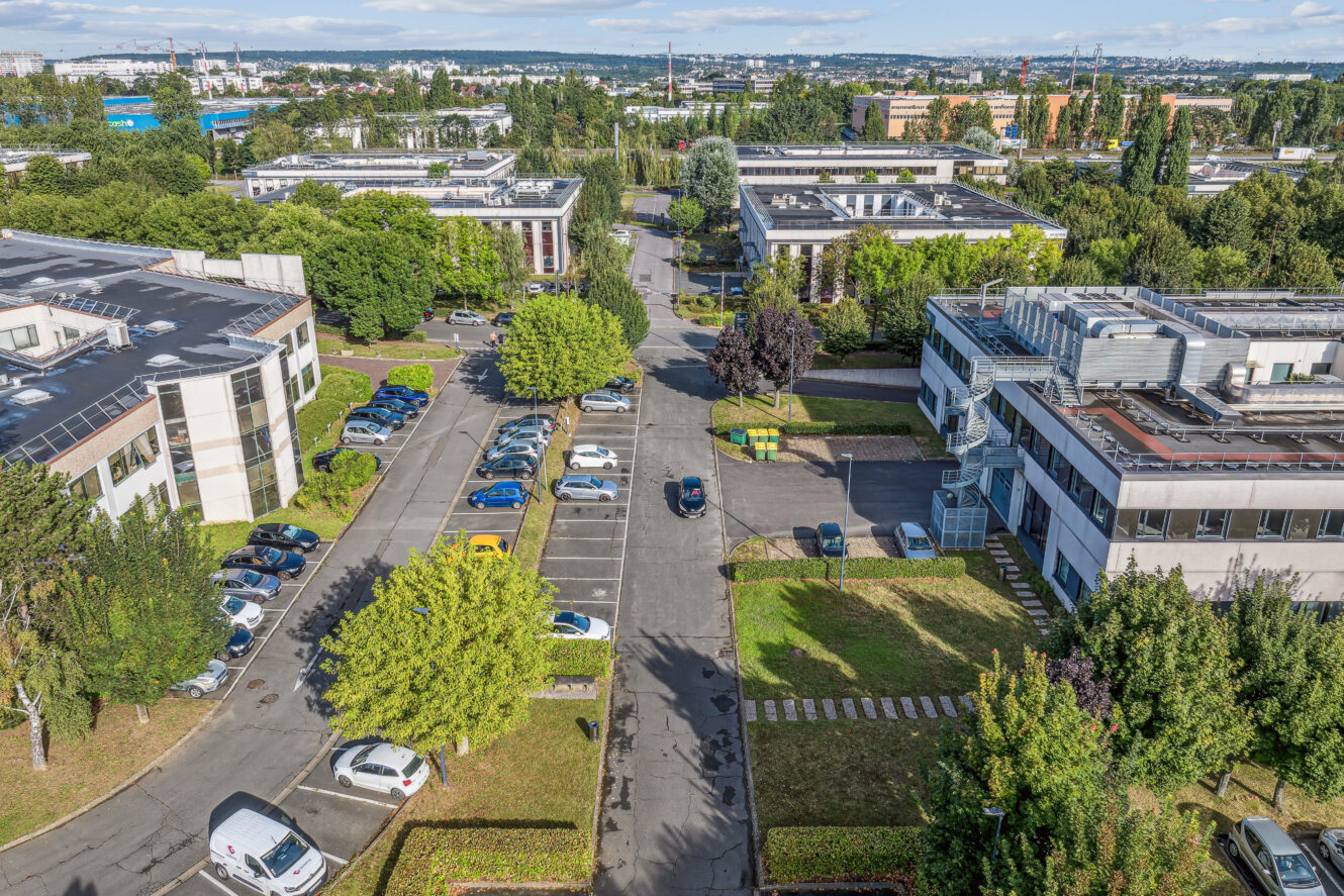 Vue aérienne d'immeubles de bureaux, de parkings et de voitures garées, entourés d'arbres, dans un parc d'activités de banlieue.