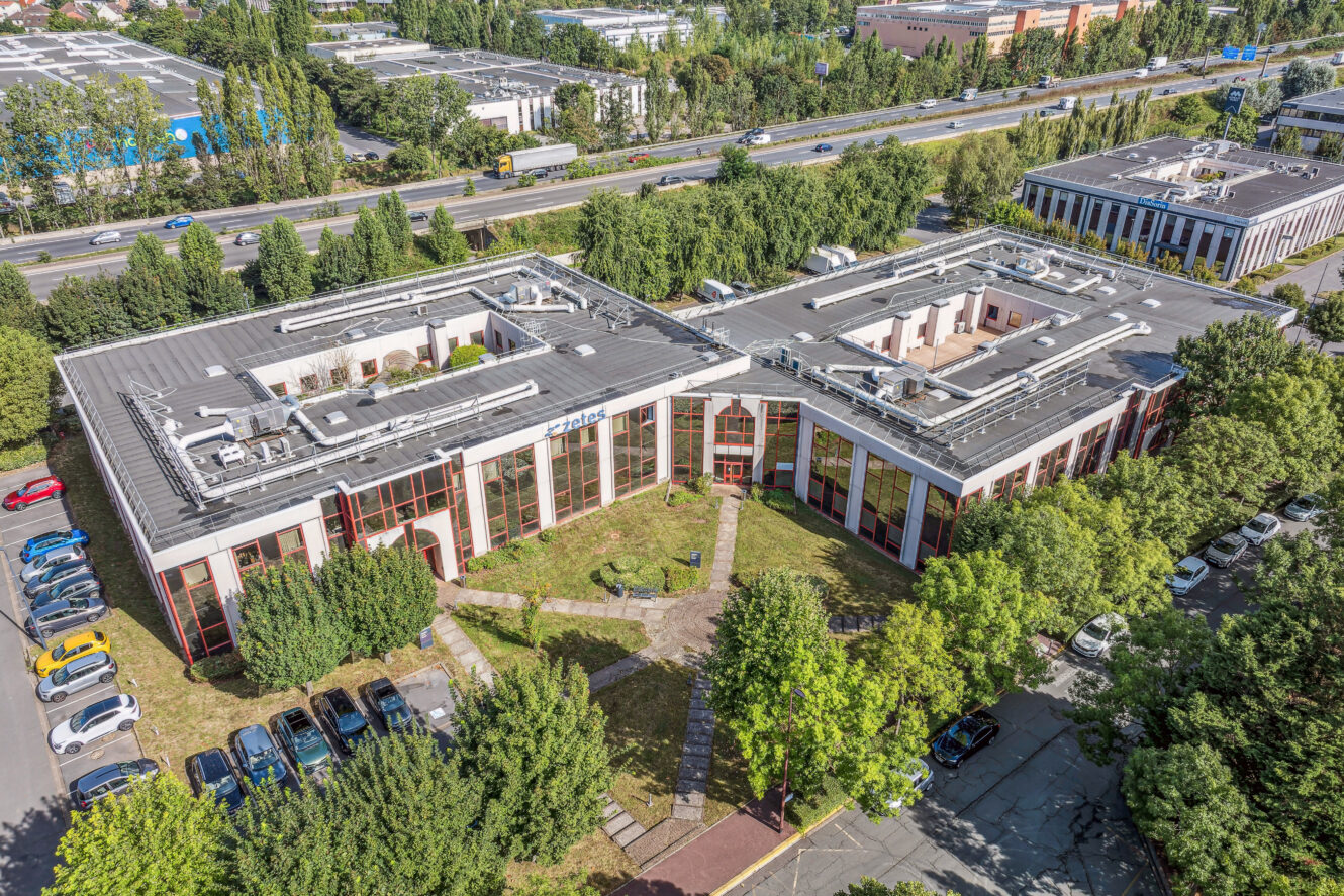 Vue aérienne d'un immeuble de bureaux moderne avec une cour centrale, entouré d'arbres, de voitures garées et adjacent à une route très fréquentée.
