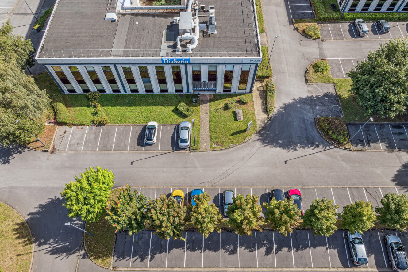 Vue aérienne d'un parking avec plusieurs voitures, d'un terre-plein bordé d'arbres et d'un bâtiment portant le nom de DiaSorin par temps clair.
