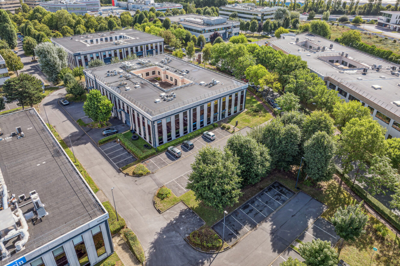Vue aérienne d'un parc d'affaires avec des immeubles de bureaux modernes, des parkings et des arbres environnants, par une journée ensoleillée.