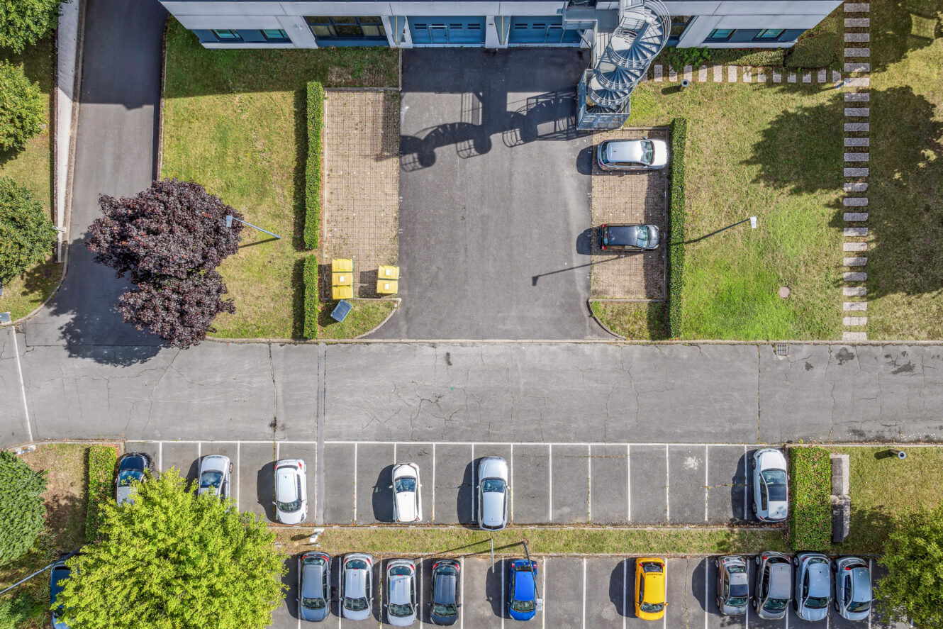 Vue aérienne d'un parking avec des voitures garées sur des places marquées, des pelouses vertes, un bâtiment au sommet et un escalier en colimaçon visible à l'extérieur du bâtiment.