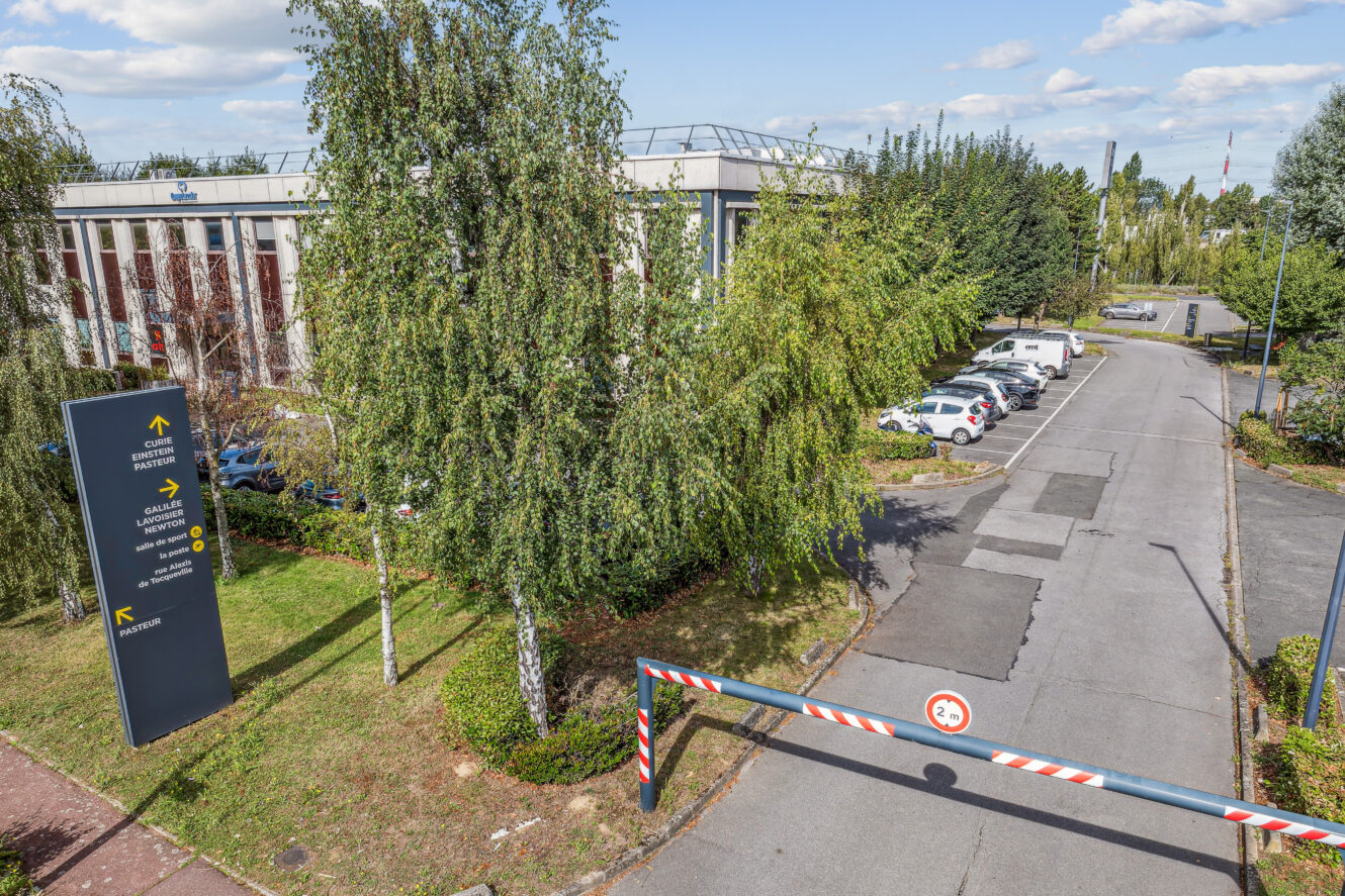 Entrée fermée d'un parc d'activités avec un panneau directionnel, des arbres, des voitures garées et un immeuble de bureaux à plusieurs étages en arrière-plan, sous un ciel partiellement nuageux.