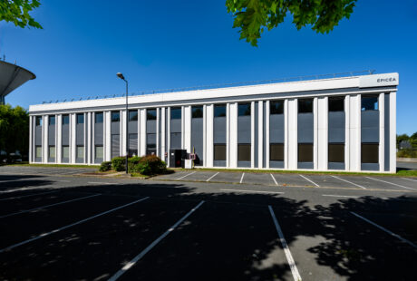 Un immeuble de bureaux de deux étages avec des colonnes verticales blanches, de grandes fenêtres et un panneau portant l'inscription EPICEA, vu d'un parking vide par temps clair.
